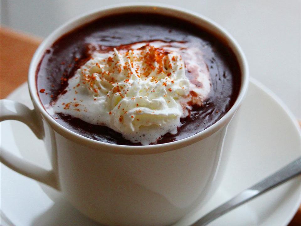 rich and creamy-looking hot chocolate topped with whipped cream and a sprinkle of spice in a white cup with a saucer