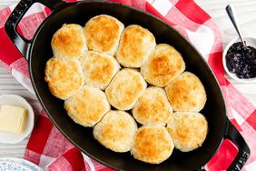 An oval cast iron pan full of golden brown biscuits on top of a red and white checkered towel with butter and jam on the sides. 