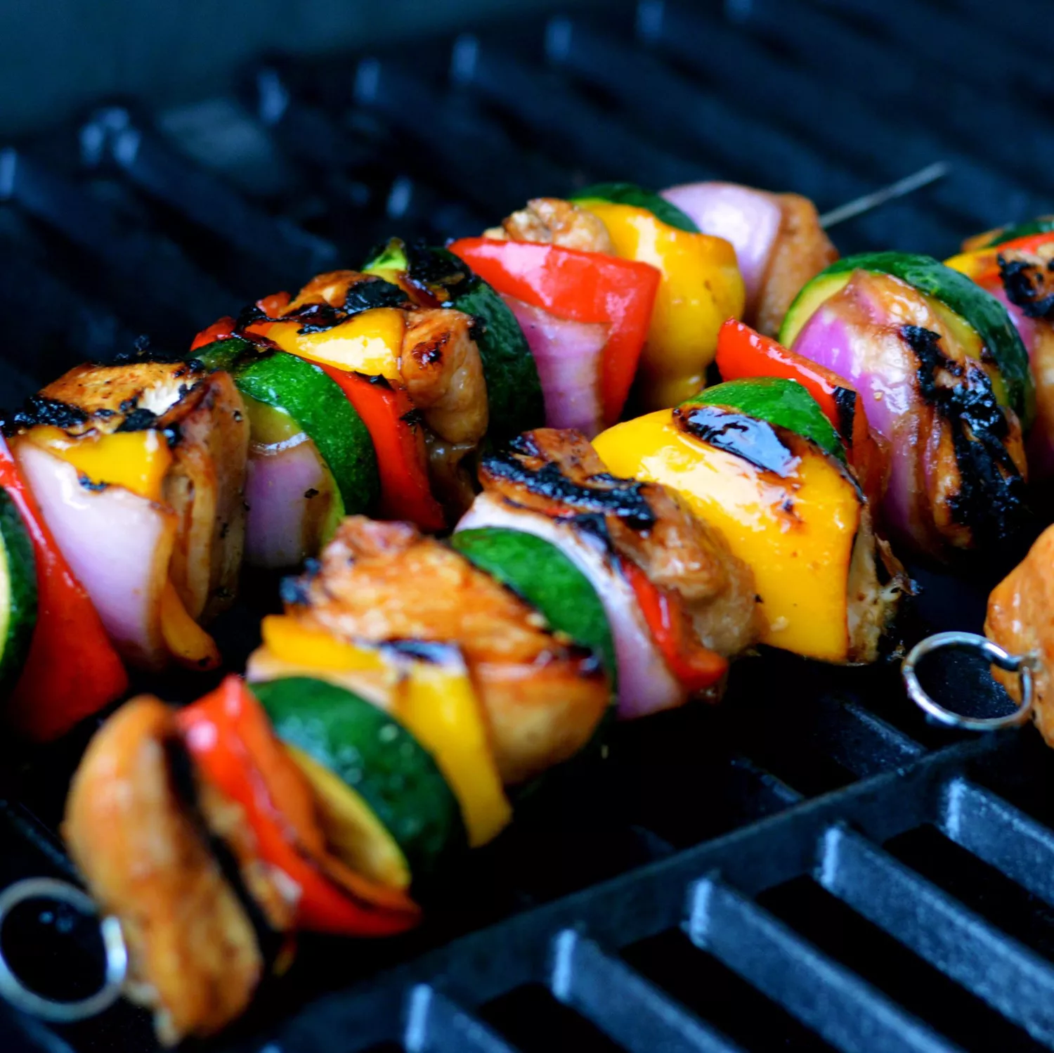 two colorful skewers of chicken, red bell pepper, zucchini, red onion, and yellow squash on a grill